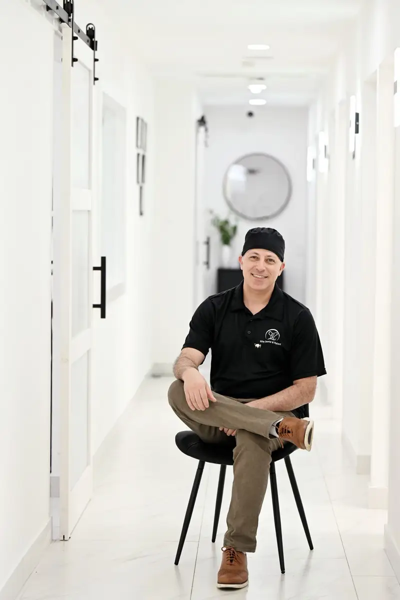 Smiling professional wearing a black shirt and cap sitting on a chair in a modern clinic hallway.